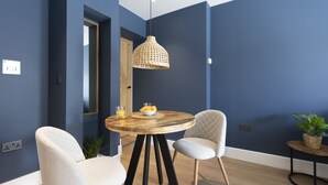 Un coin repas comprenant une table ronde en bois, deux chaises, un luminaire suspendu en fibres tressées et une petite table d’appoint agrémentée d’une plante.