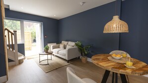 A modern living room with a sofa, a dining table with a bowl of fruit and a glass of orange juice, and a hanging light fixture. - Bell House Oxford - The Noke (Oxford)