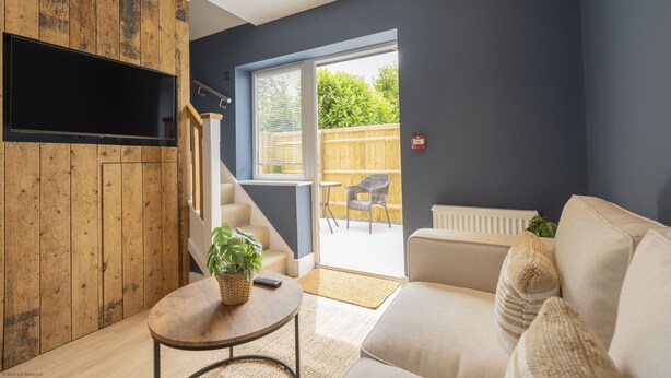 A modern living room with a sofa, a coffee table, and a TV mounted on a wooden wall. There is a sliding glass door leading to an outdoor area with a chair and a plant. - Bell House Oxford - The Noke (Oxford)