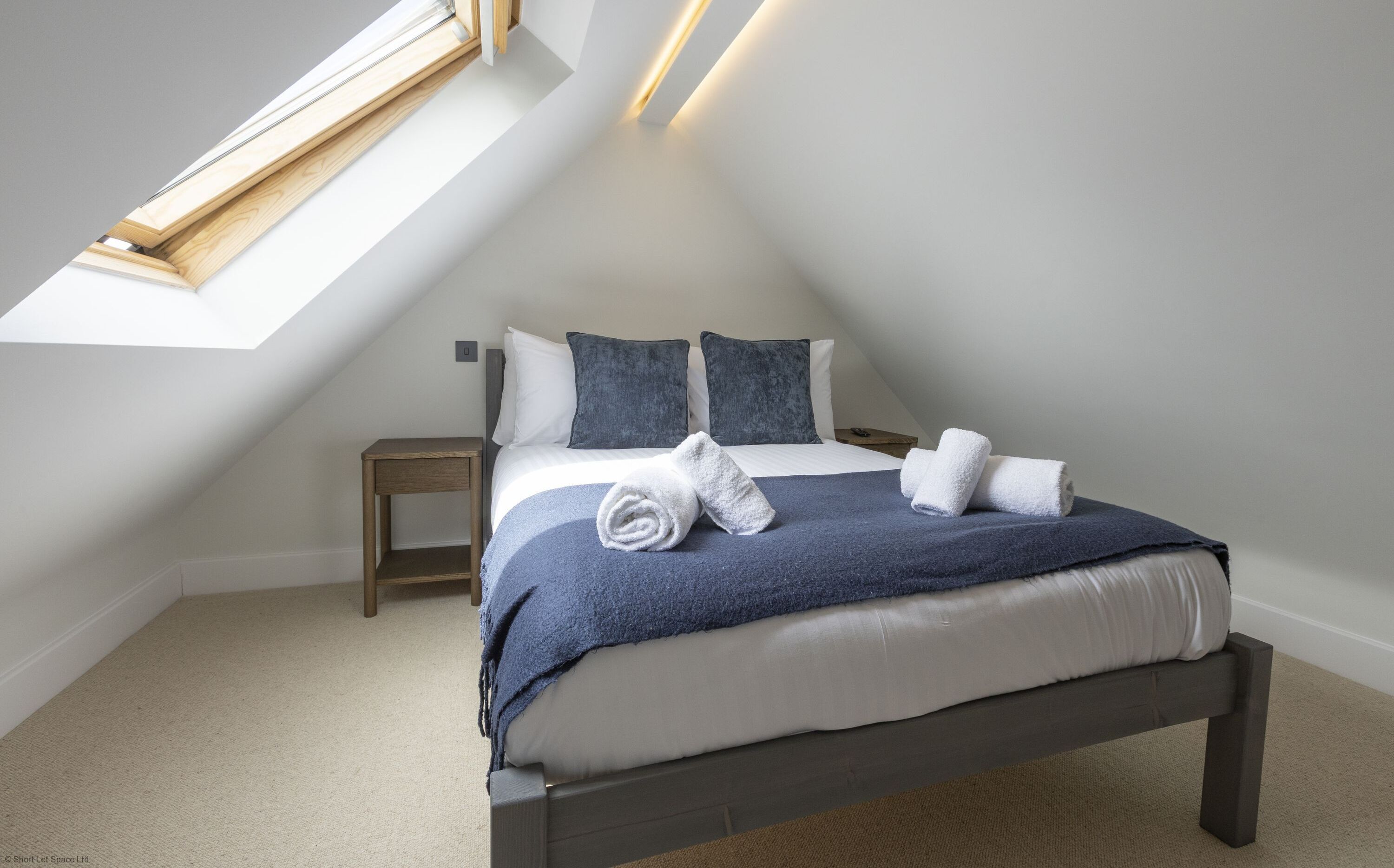 A bedroom with a sloped ceiling, a skylight, a bed with a blue blanket, two pillows, and two rolled towels on the bed.