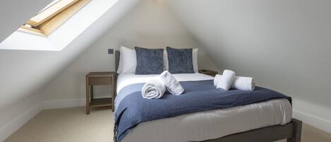 A bedroom with a sloped ceiling, a skylight, a bed with a blue blanket, two pillows, and two rolled towels on the bed.