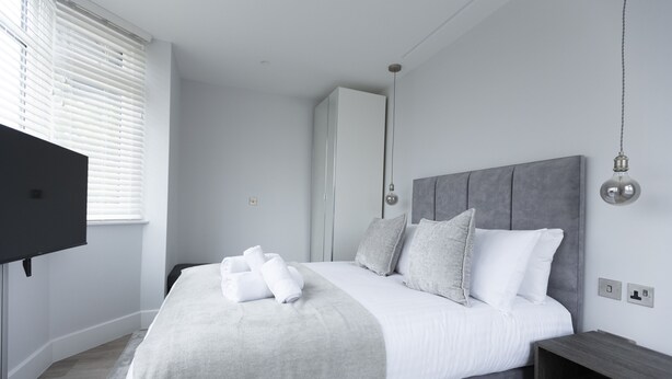 A modern bedroom with a large bed, grey bedding, a flat-screen TV, and a wardrobe. - Bell House Oxford - The Punt (Oxford)