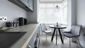 A modern kitchen with a dining area, featuring a round table, two chairs, and a window with blinds. - Bell House Oxford - The Punt (Oxford)