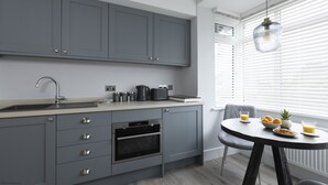 A modern kitchen with grey cabinets, a built-in oven, a sink, and a dining area with a table and chairs. - Bell House Oxford - The Punt (Oxford)