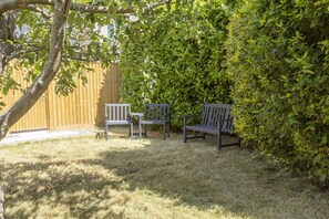 Un espace patio avec deux bancs en bois et une petite table, entouré d’un feuillage verdoyant et dense et d’une clôture en bois.