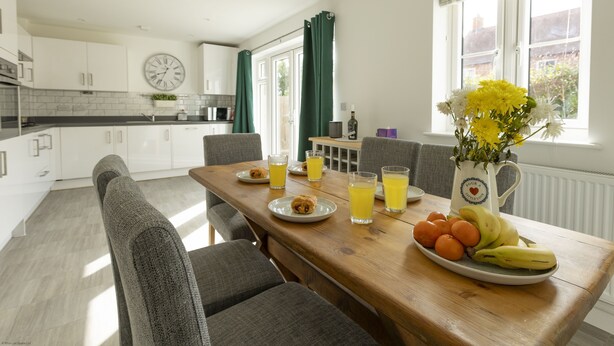 A dining area with a wooden table set for breakfast, including plates of fruit, glasses of juice, and a pitcher of flowers. - Wren House in Bicester (Bicester)