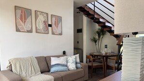 A modern living room with a sofa, a coffee table, and a dining area with a table and chairs. There are abstract paintings on the wall and a staircase leading to an upper level.