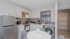 Cocina compacta con refrigerador, microondas, estufa, pileta y lavadora. Hay una mesa de comedor con sillas y una ventana con persianas.