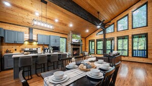 A spacious kitchen with a central island, a dining area with a table set for four, and large windows offering views of the surrounding forest. - Timberline Pines · Spacious Escape | Hot Tub, Fireplaces & Soaking Tub (Broken Bow)