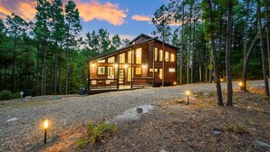 A two-story log cabin with a deck, surrounded by a forest. - Timberline Pines · Spacious Escape | Hot Tub, Fireplaces & Soaking Tub (Broken Bow)