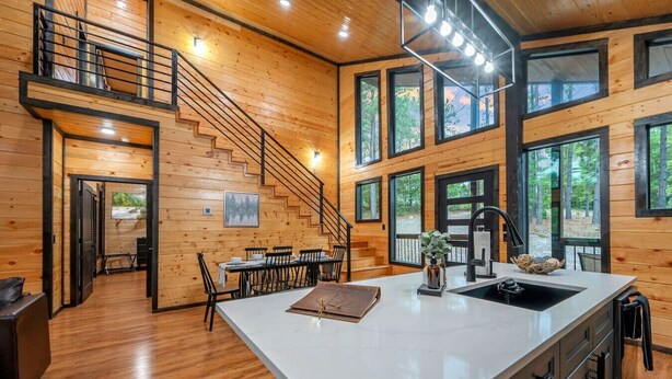 A spacious cabin interior with a large kitchen island, wooden walls, and a staircase leading to a loft area. - Timberline Pines · Spacious Escape | Hot Tub, Fireplaces & Soaking Tub (Broken Bow)