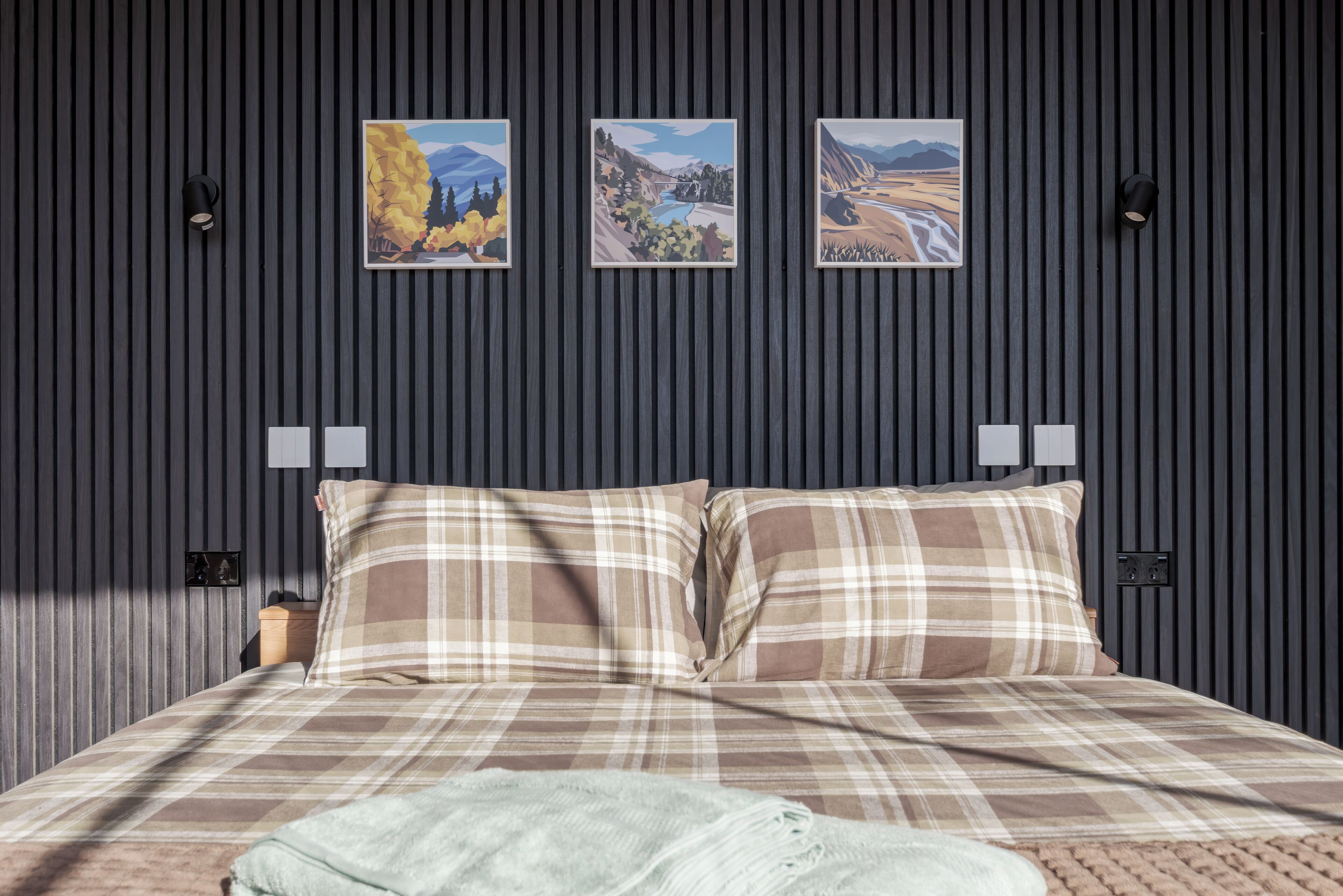A bed with plaid bedding, a light throw blanket, and three framed landscape paintings on a dark striped wall.