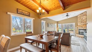 A dining area with a wooden table and chairs, a chandelier, and a view of the outdoors through large windows. - NEW! All Season Hot Tub, Fire Pit, Golf Course, Breezy Point (Pequot Lakes)
