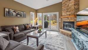 A living room with a stone fireplace, a ceiling fan, a large window, and a couch. - NEW! All Season Hot Tub, Fire Pit, Golf Course, Breezy Point (Pequot Lakes)
