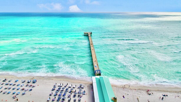 Ein Strand mit einer langen Seebrücke, die sich in das türkisblaue Meer erstreckt, sowie Strandsesseln und Sonnenschirmen im Sand.