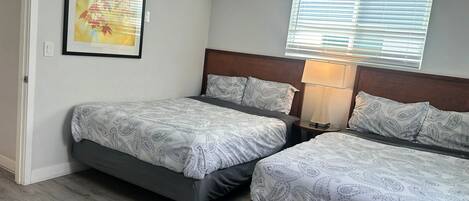 Een hotelkamer met twee bedden, een nachtkastje met lamp, een lijst met afbeelding aan de muur en een raam met zonwering.