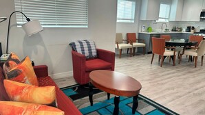 A modern living room with a red sofa, a coffee table, and a patterned rug. A dining area with wooden chairs and a kitchen in the background.