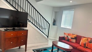A living room with a staircase, a flat-screen TV on a wooden cabinet, a red sofa with colorful cushions, and a blue patterned rug.