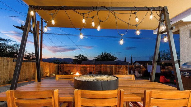 An outdoor patio with a round table and chairs, overhead string lights, and a fire pit. - Cozy Cactus - Cowboy Pool, Outdoor Movies, 3BR - Walk to Saloon (Joshua Tree)