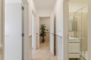 Un couloir étroit avec un pot de plante, une cabine de douche en verre et un lavabo blanc.
