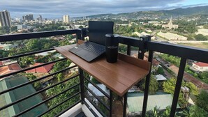 A balcony with a laptop and a tumbler on a wooden table, overlooking a cityscape with buildings and greenery. - Studio Unit with Balcony + City & Mountain Views + Fast WiFi | Near IT Park (Cebu City)