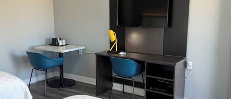 A hotel room with a bed, a desk with two chairs, a flat-screen TV mounted on the wall, and a small table with a coffee maker and salt and pepper shakers.