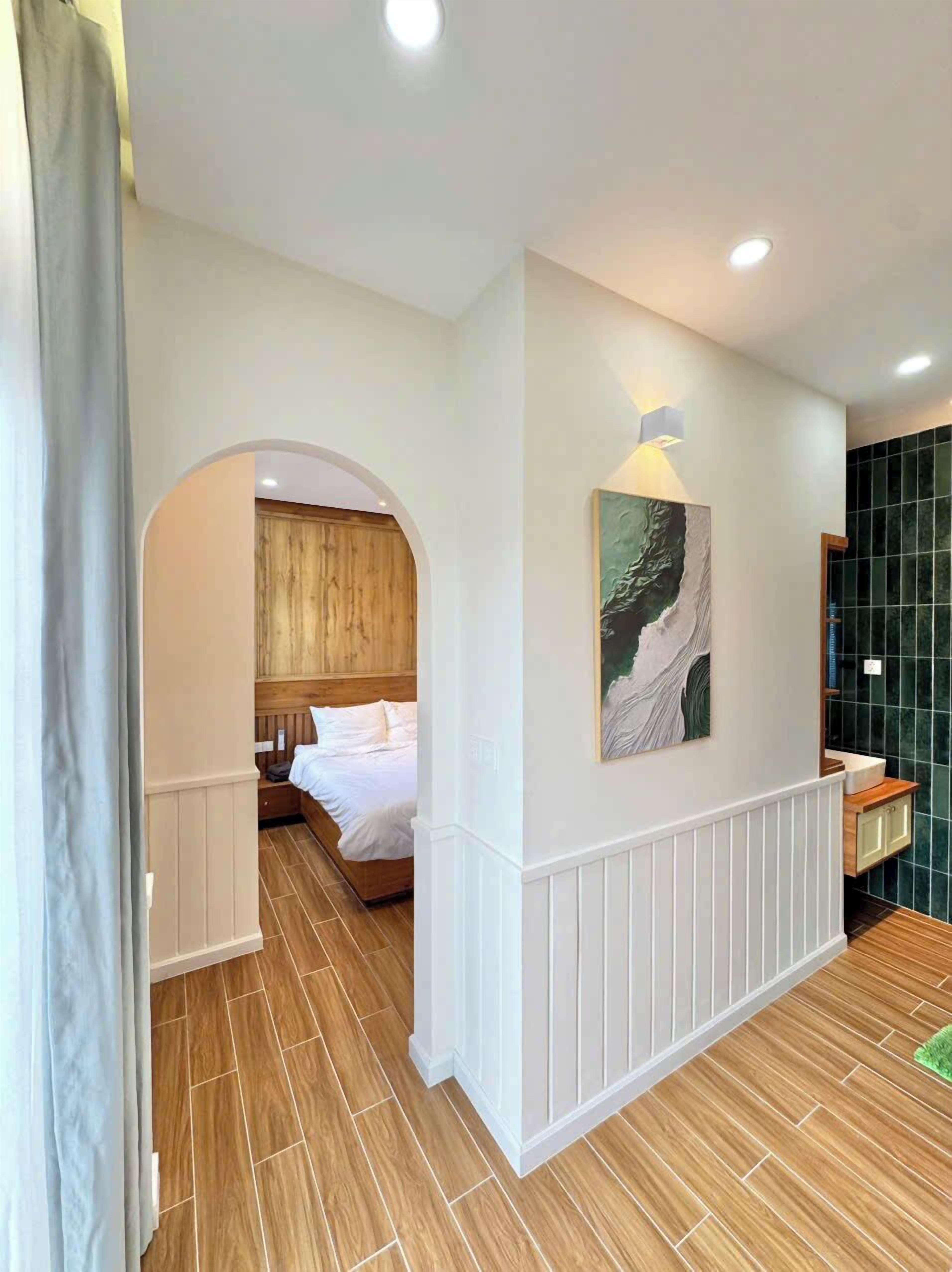 Habitación de hotel con suelo de madera, cama con ropa blanca, cabecero de madera, un cuadro en la pared y un muro con azulejos verdes.