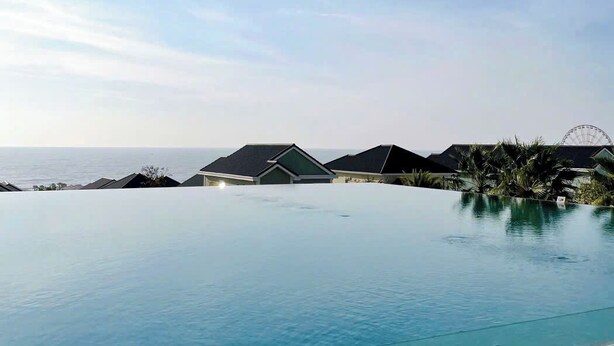 Un cielo azul despejado con una amplia piscina infinita, una noria y varios edificios con techos oscuros al fondo.