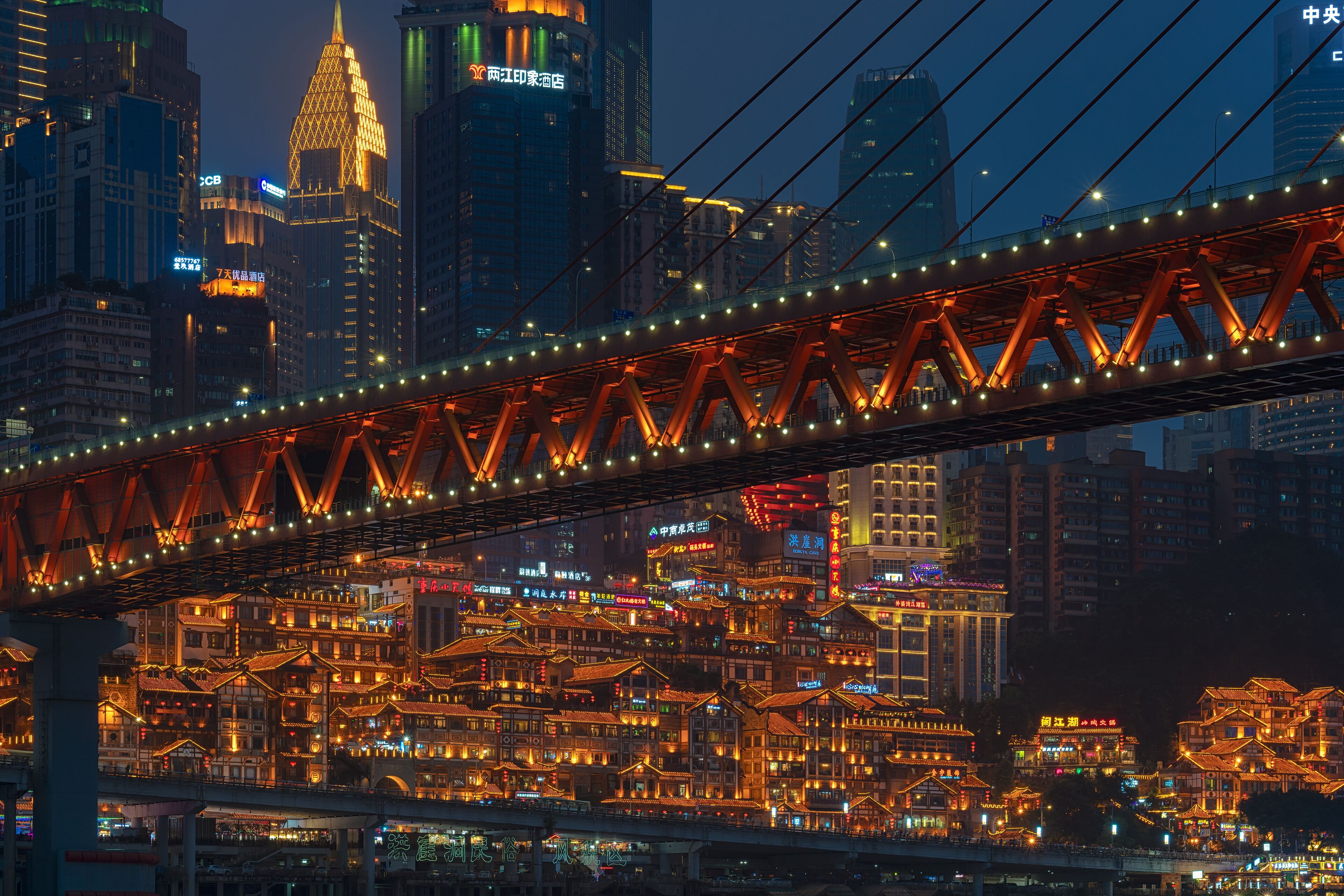 A bridge with illuminated lights spanning over a densely packed urban area with traditional buildings and modern skyscrapers.