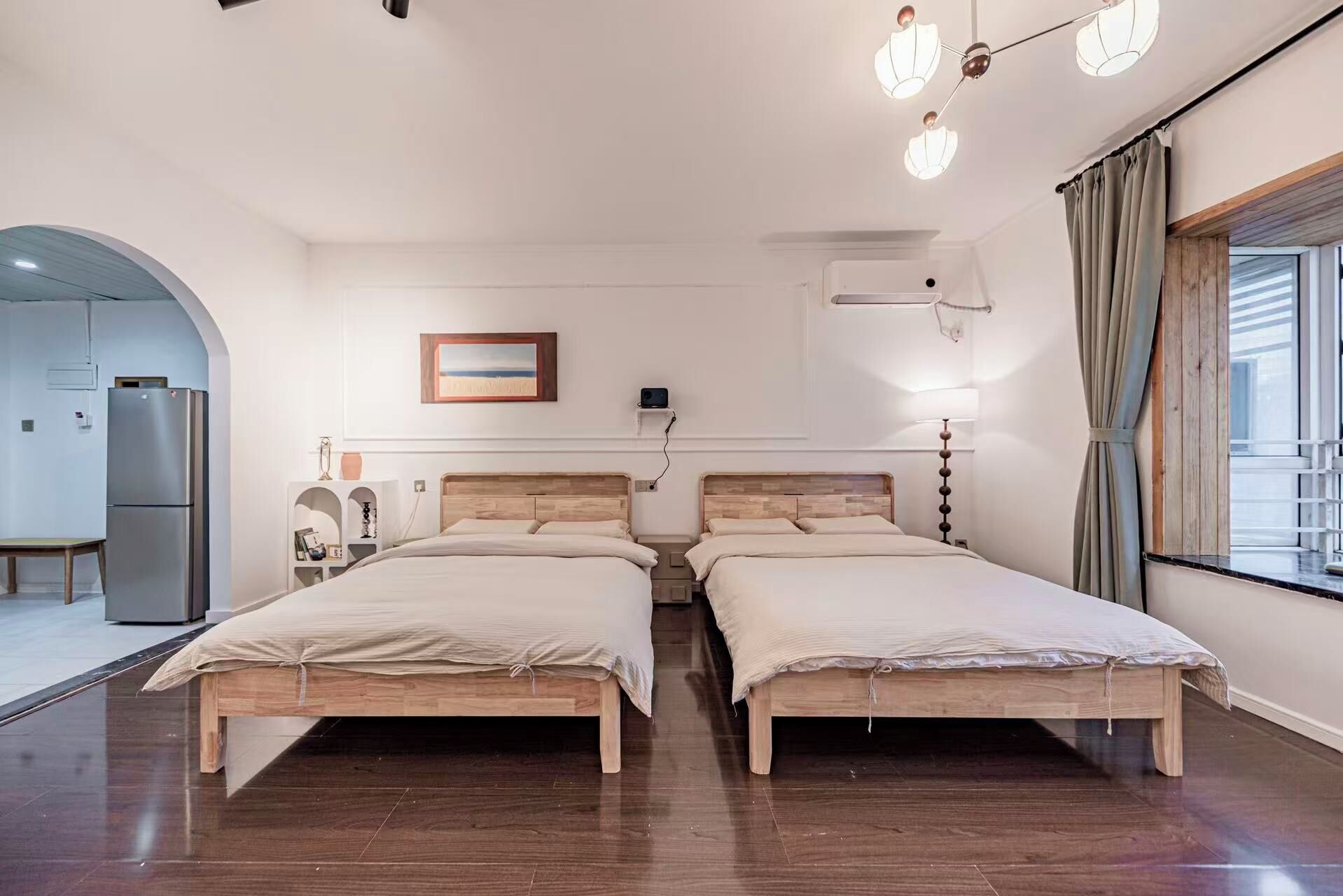 Habitación de hotel con dos camas de madera, una mesita, un refrigerador, un cuadro en la pared y una ventana con cortinas.