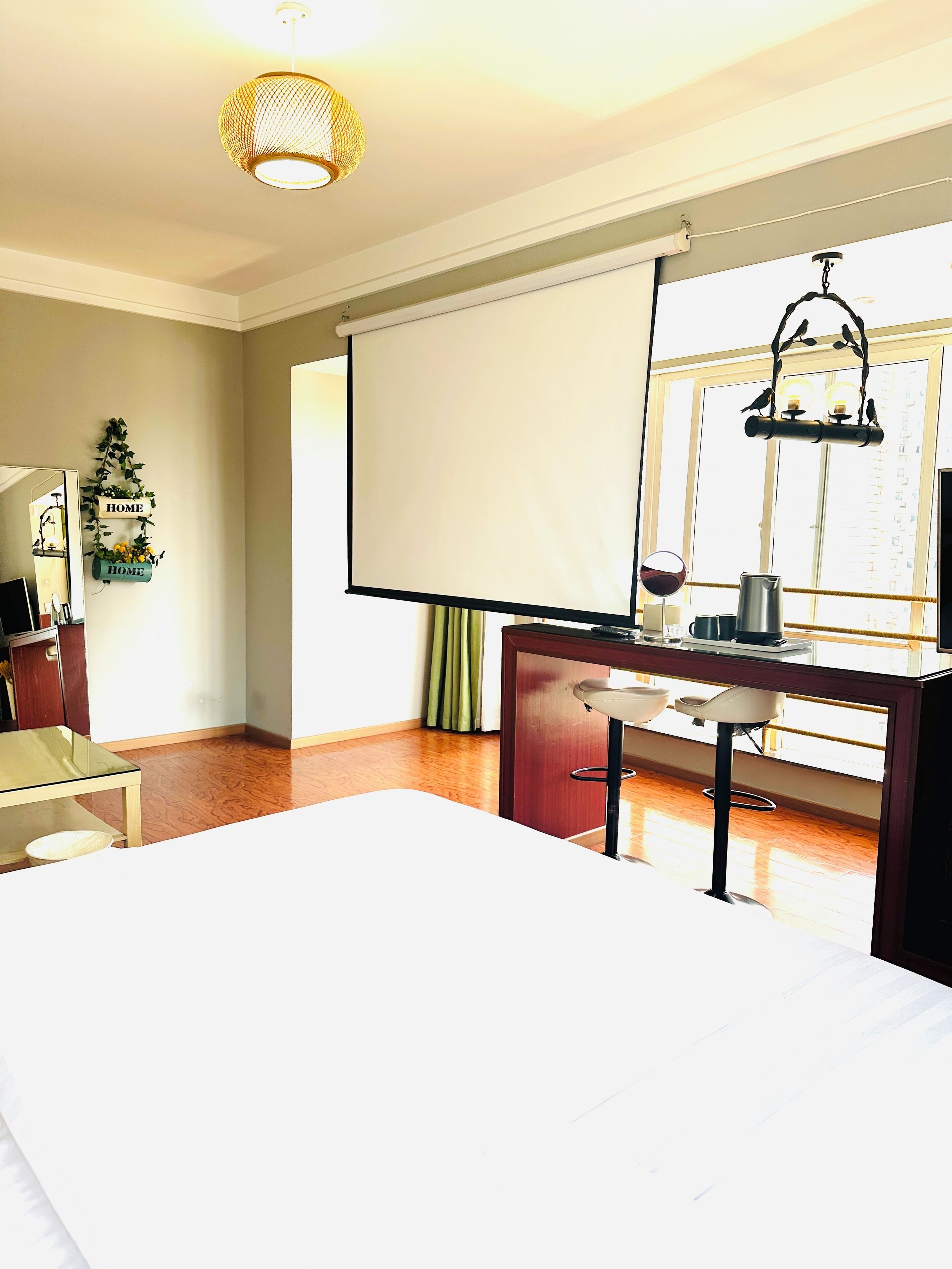 A hotel room with a bed, a desk with a chair, a large window, and a projector screen.