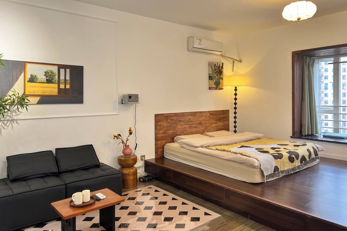 Un dormitorio moderno con una cama de madera con cabecera, un sofá negro, una mesita con dos tazas, una lámpara y un cuadro en la pared.