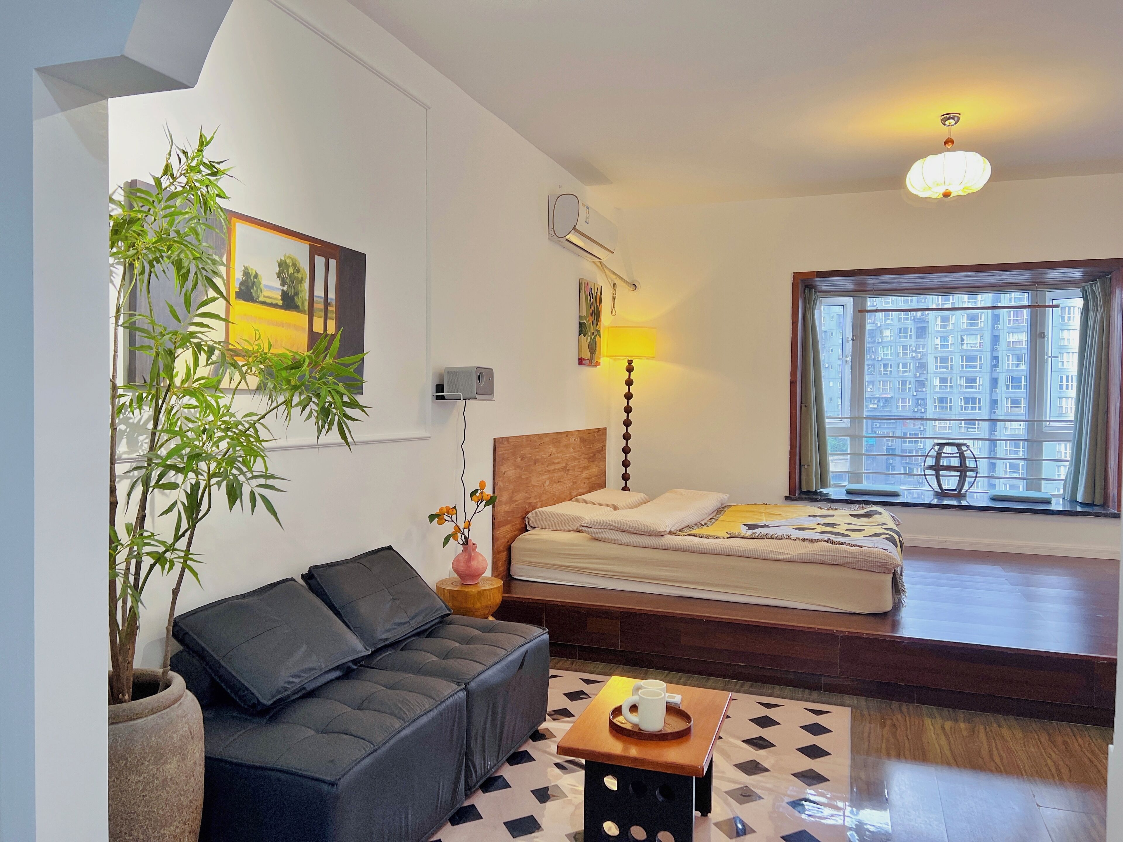 Un dormitorio moderno con cama, un sofá, una mesita, una planta en maceta y una ventana con vistas a la ciudad.