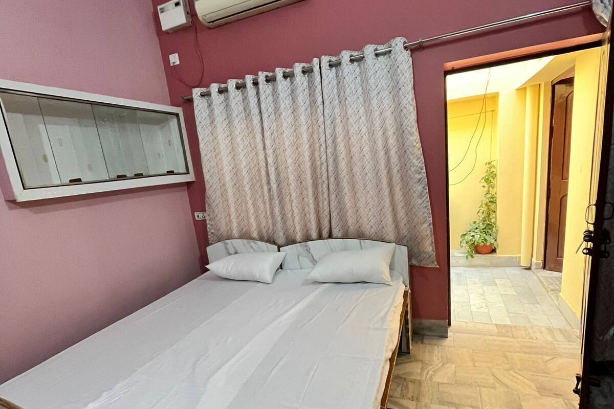 A bedroom with a single bed, an air conditioning unit, a mirror, and a view of a hallway with a door leading outside.