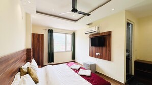 A hotel room with a bed, a ceiling fan, an air conditioning unit, a television mounted on the wall, and a window with curtains. - Hotel Hitam (Rishikesh)