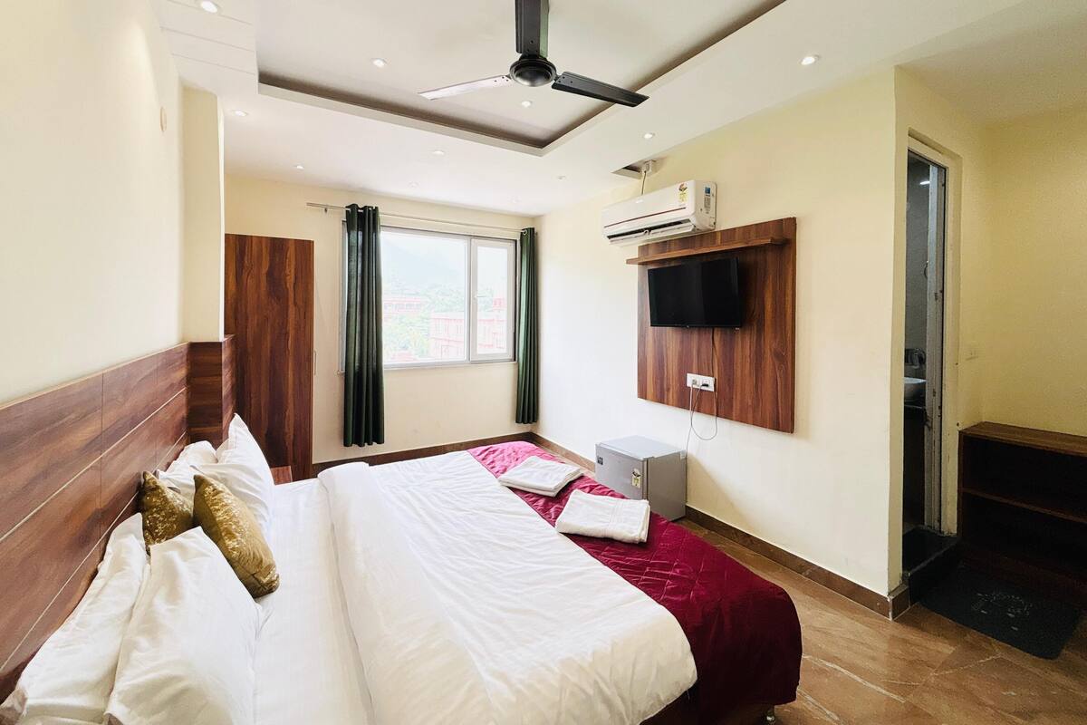 A hotel room with a bed, a ceiling fan, an air conditioning unit, a television mounted on the wall, and a window with curtains.