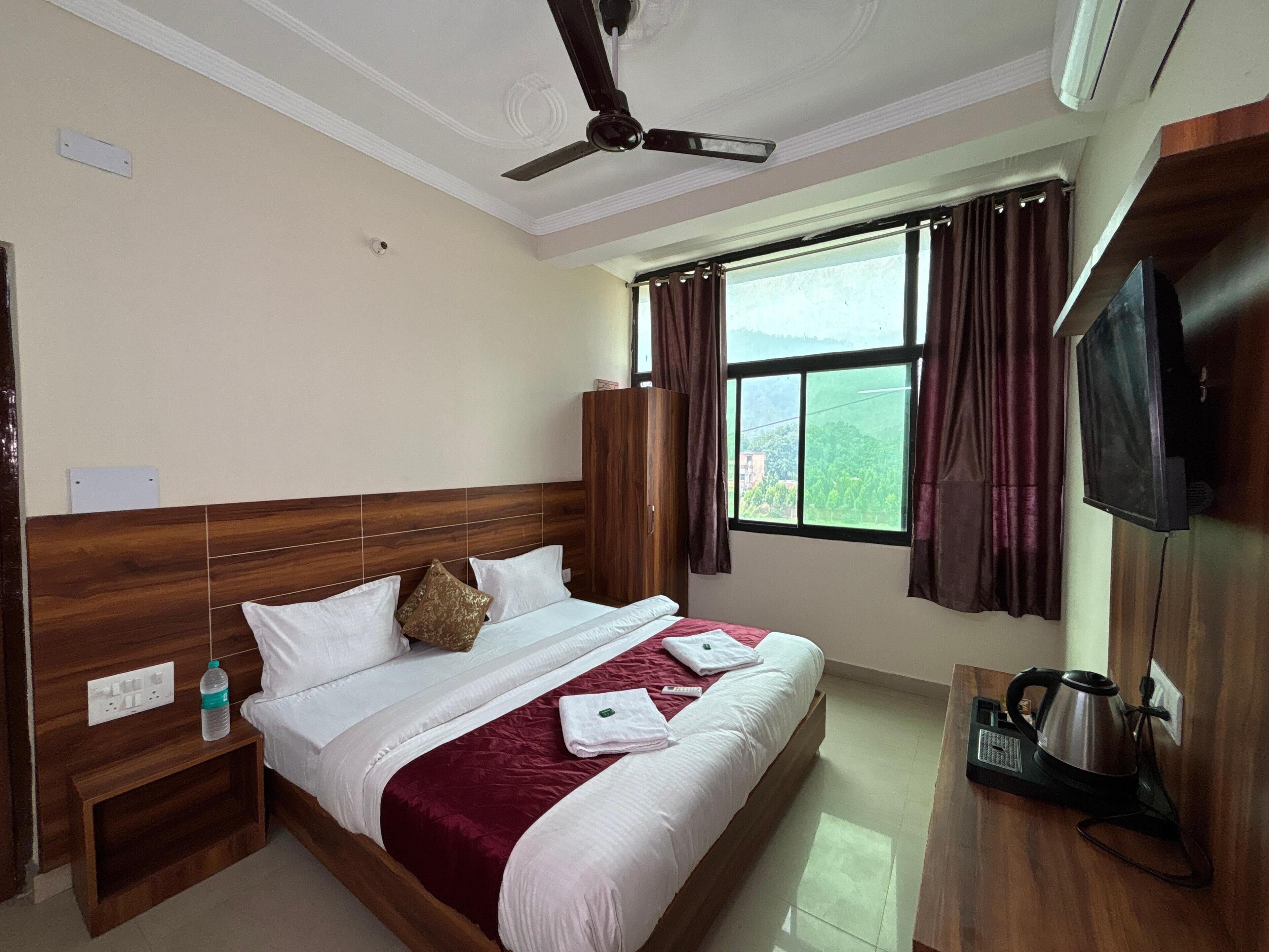 Una habitación de hotel con cabecera de madera, cama con ropa blanca y roja, ventilador de techo, televisor y ventana con cortinas.