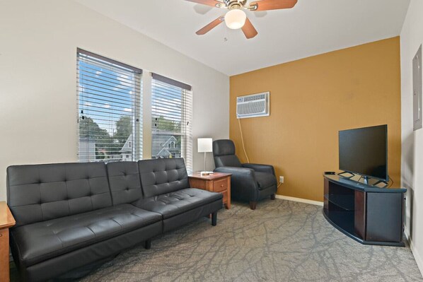 A living room with a black leather sofa, a wooden side table, a black armchair, a ceiling fan, and a television on a stand. - Maple Manor City Scene (Spokane)