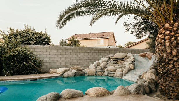 A swimming pool with a rock waterfall, surrounded by a brick wall and palm trees. - Spacious 4BDRM with a Stunning Pool+Game Room and Firepit (Bakersfield)