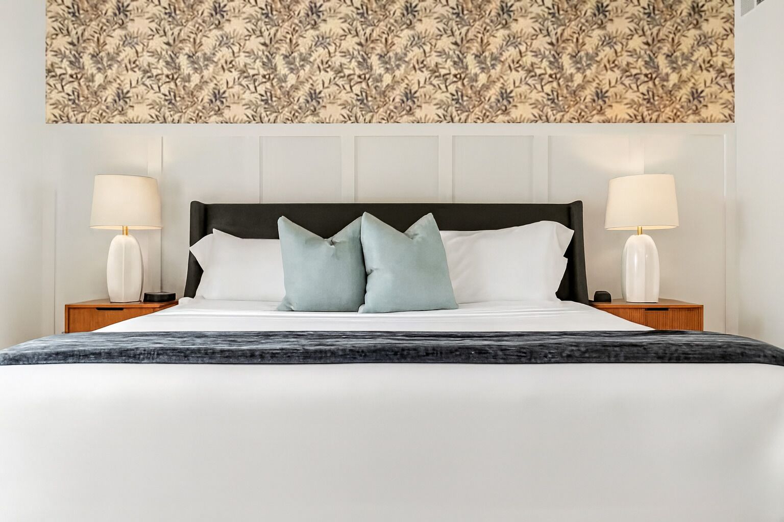 A neatly made bed with a dark headboard, two bedside lamps, and a floral-patterned wallpaper above.