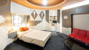 Habitación de hotel con cama, sofá rojo, escritorio con ordenador y pequeña cocina.