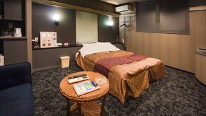 Habitación de hotel con una cama, una mesita con un control remoto y un libro, un minibar, un espejo y una ventana con persianas.