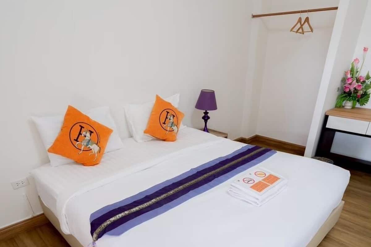 Una cama bien hecha con sábanas blancas, dos almohadas naranjas con diseño de león, una lámpara morada y una mesita de noche con un jarrón de flores.