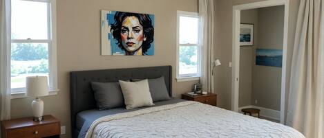 Une chambre à coucher avec un grand lit, une table de chevet, une commode, une fenêtre et un tableau accroché au mur.
