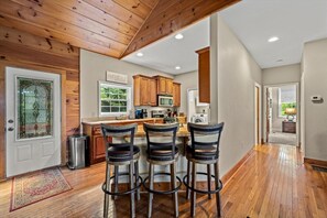 A kitchen with wooden cabinets, a central island with bar stools, and a view of the outdoors through a window. - Secluded Mountain Top with Game Room and Fire Pit Near Boone (Boone)
