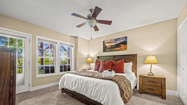 A bedroom with a bed, two bedside lamps, a ceiling fan, and a window with a view of trees.