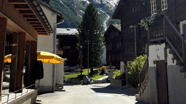 A mountain village street with traditional wooden buildings, a clear sky, and a snow-capped mountain in the background. - Haus Salü 5 by Interhome (Saas-Fee)