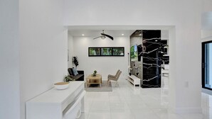 Un salon moderne et bien éclairé, avec une console blanche, un canapé, une table basse et un fauteuil. On y trouve également un ventilateur de plafond, un tableau encadré au mur et un mur d’accent en marbre noir.