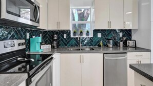 A modern kitchen with a microwave, coffee maker, and toaster. There is a window with plants and a view of trees. The kitchen has white cabinets and a black countertop. - Palm Breeze Retreat . Close to Beach & Downtown (Hollywood)