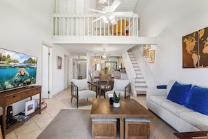 Een ruime woonkamer met een hoge plafond, een balkon, een eethoek en een bank met blauwe kussens.